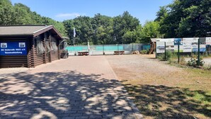 Una cabana de fusta amb un senyal que indica que és una instal·lació de tennis, un camí pavimentat que hi porta, i una zona tancada amb bancs i un vèrtex.