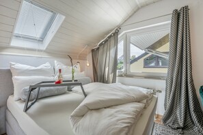 A bedroom with a bed, a bedside table with a bottle and glasses, a window with curtains, and a skylight.