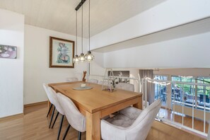 A dining area with a wooden table, cushioned chairs, a hanging light fixture, and a framed artwork on the wall.