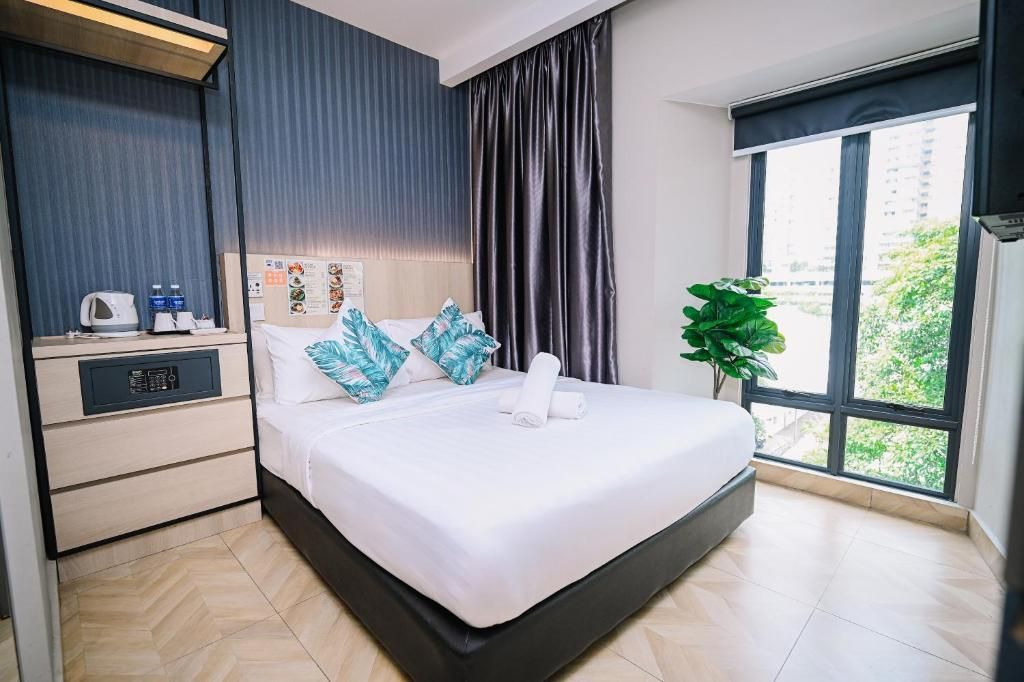 Habitación de hotel moderna con una cama grande, una mesita de noche con hervidor y botellas, una planta y una ventana con vistas a la ciudad.