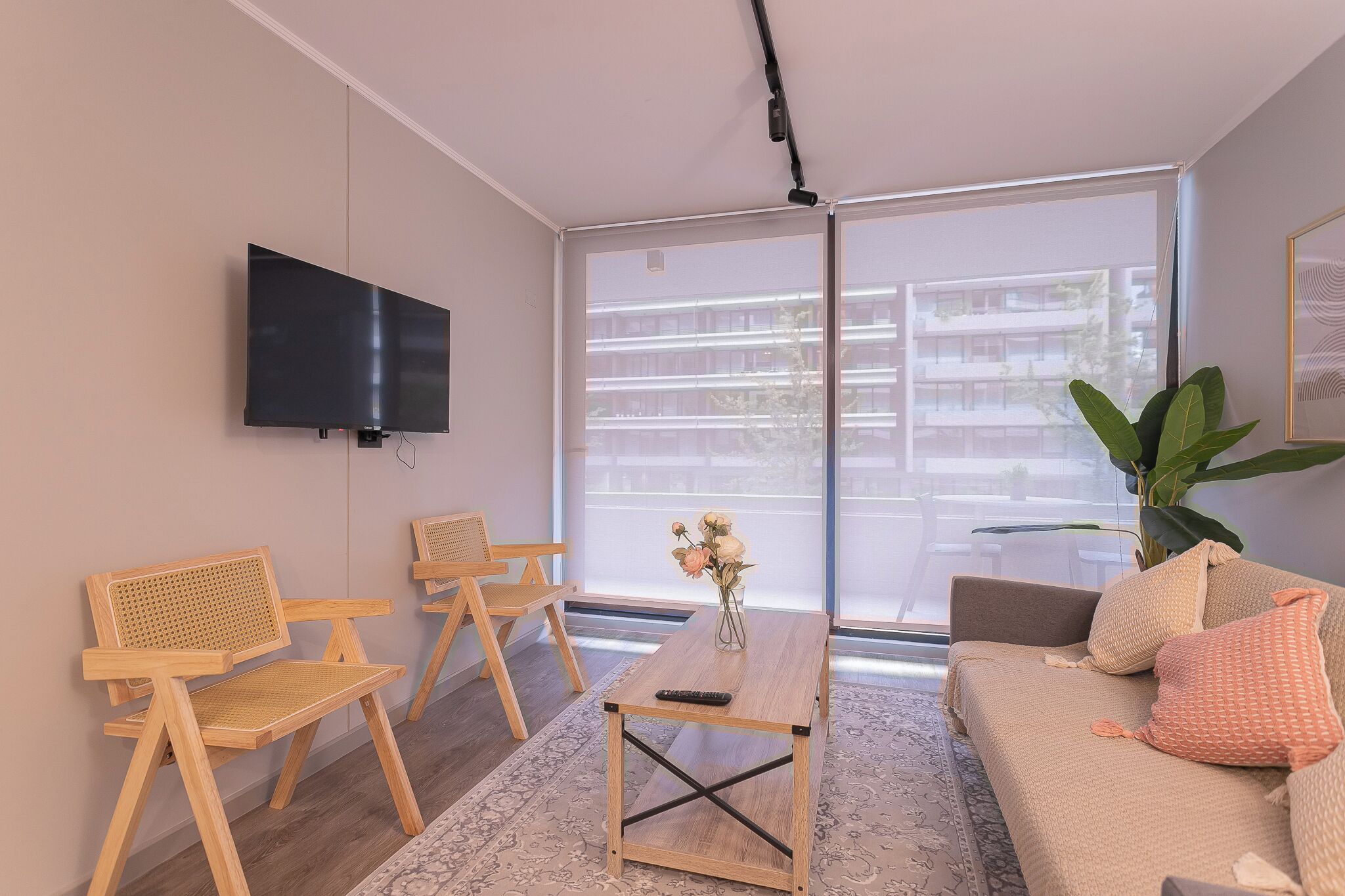 Een moderne woonkamer met een flatscreen-televisie aan de wand, een bank, twee houten stoelen, een salontafel en een groot raam met uitzicht op een gebouw.