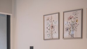 Un dormitorio con una cama, dos almohadas, una mesita de noche con una lámpara y dos cuadros con motivos florales en la pared.