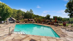 A rectangular swimming pool with a stone deck, surrounded by lounge chairs and umbrellas, with buildings and trees in the background. - Luxury Lakefront Condo, Gated Community, Mountain & Lake Views, Pool, Dock, & Kayaks (Lake Lure)