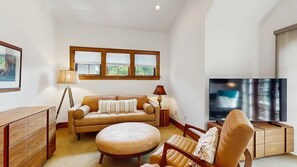 Ein Wohnzimmer mit Sofa, Hocker, Beistelltisch, Lampe, Fernseher und Holzschrank.