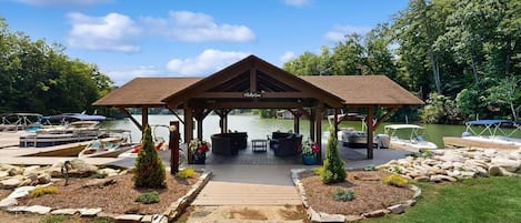 A covered pavilion with a wooden roof, a stone pathway leading to it, and a dock with boats.