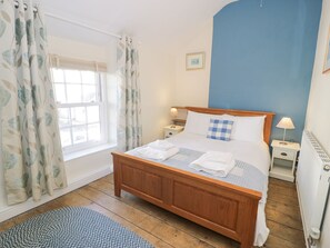 Ein Schlafzimmer mit einem hölzernen Bett, weißer Bettwäsche, einer blauen Wand, einem Fenster mit Vorhängen, einem Nachttisch mit Lampe und einem Heizkörper.
