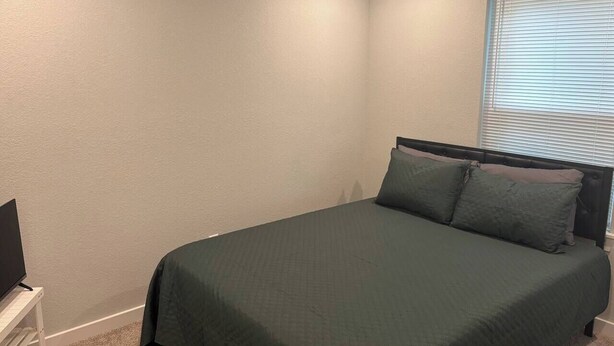 A bedroom with a bed, a television on a stand, and a window with blinds. - Cozy and spacious home 15 minutes from downtown. (Houston)
