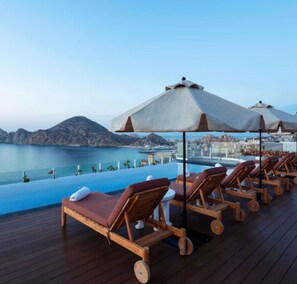 Eine Terrasse mit Liegestühlen und Sonnenschirmen mit Blick auf ein Gewässer und eine Berglandschaft.