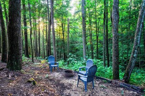Uma floresta com duas cadeiras Adirondack e uma fogueira.