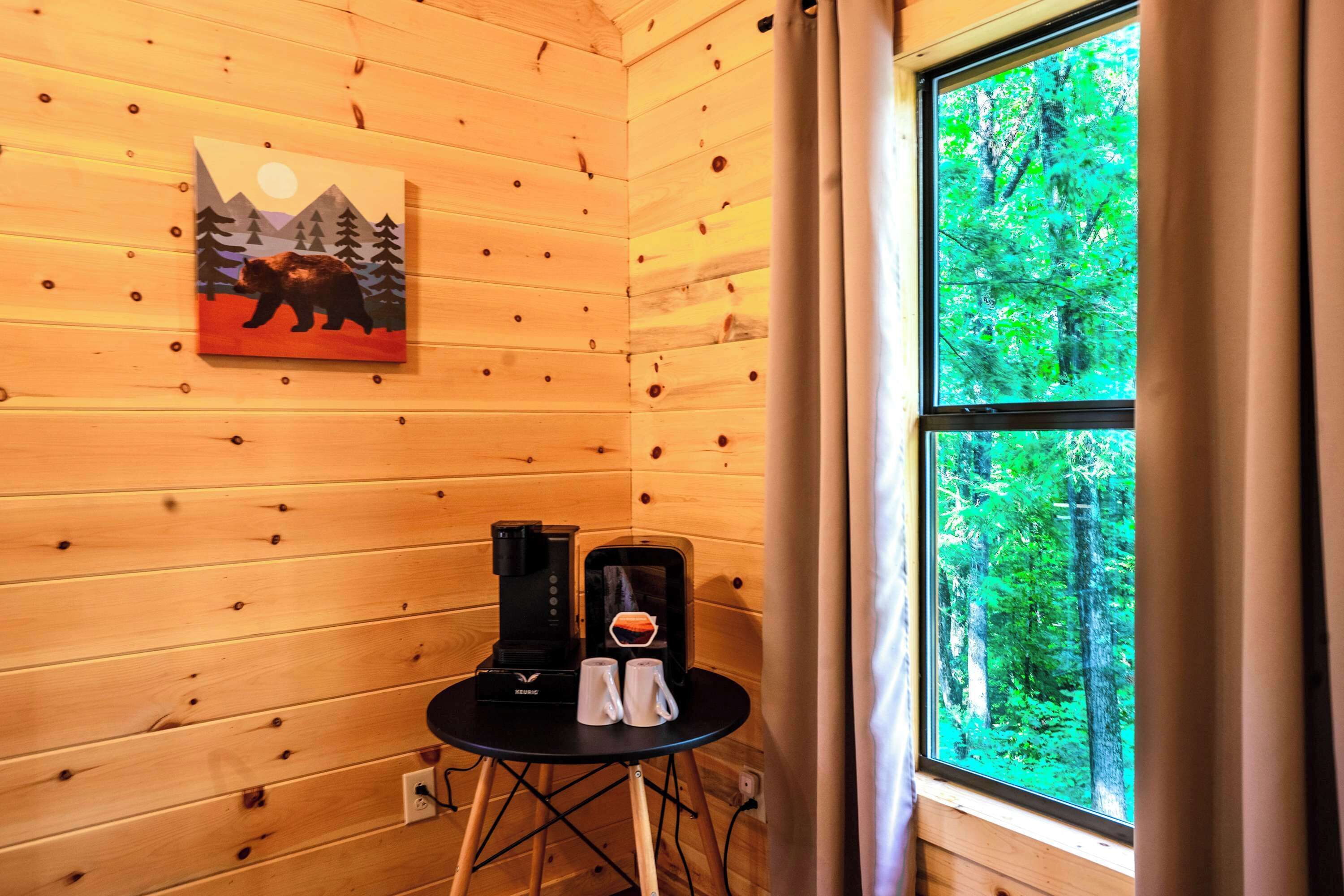 Interior de una cabaña de madera con un cuadro de un oso y montañas, una cafetera y una ventana con vistas a un bosque.