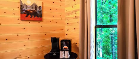 Interior de una cabaña de madera con un cuadro de un oso y montañas, una cafetera y una ventana con vistas a un bosque.