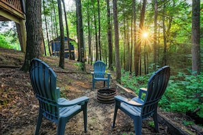 Ein bewaldetes Gelände mit zwei Adirondack-Stühlen, die sich gegenüberliegen, einer Feuerstelle in der Mitte und einer Holzhütte im Hintergrund.