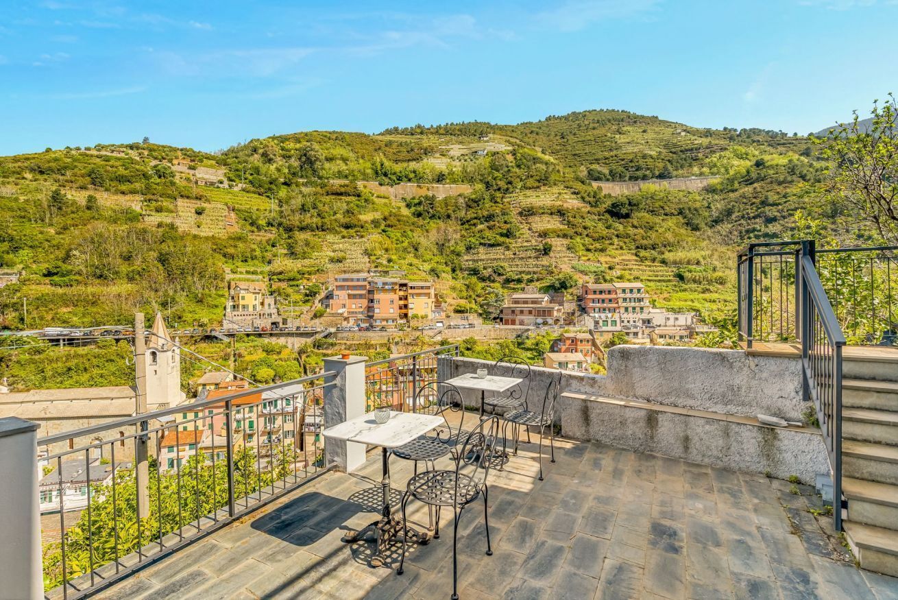 A terrace with outdoor furniture, a view of a hillside village, and a stone staircase.