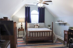 A bedroom with a sloped ceiling, a bed with a wooden headboard, a window with curtains, a lamp, and a small shelf.