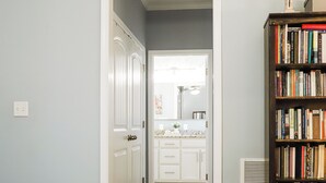 Un pasillo que conduce a un baño con una puerta blanca, un espejo y una encimera con un lavamanos. Se ve una estantería llena de libros a la derecha.