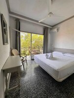 Habitación de hotel con cama, escritorio, silla y balcón con vistas a árboles.