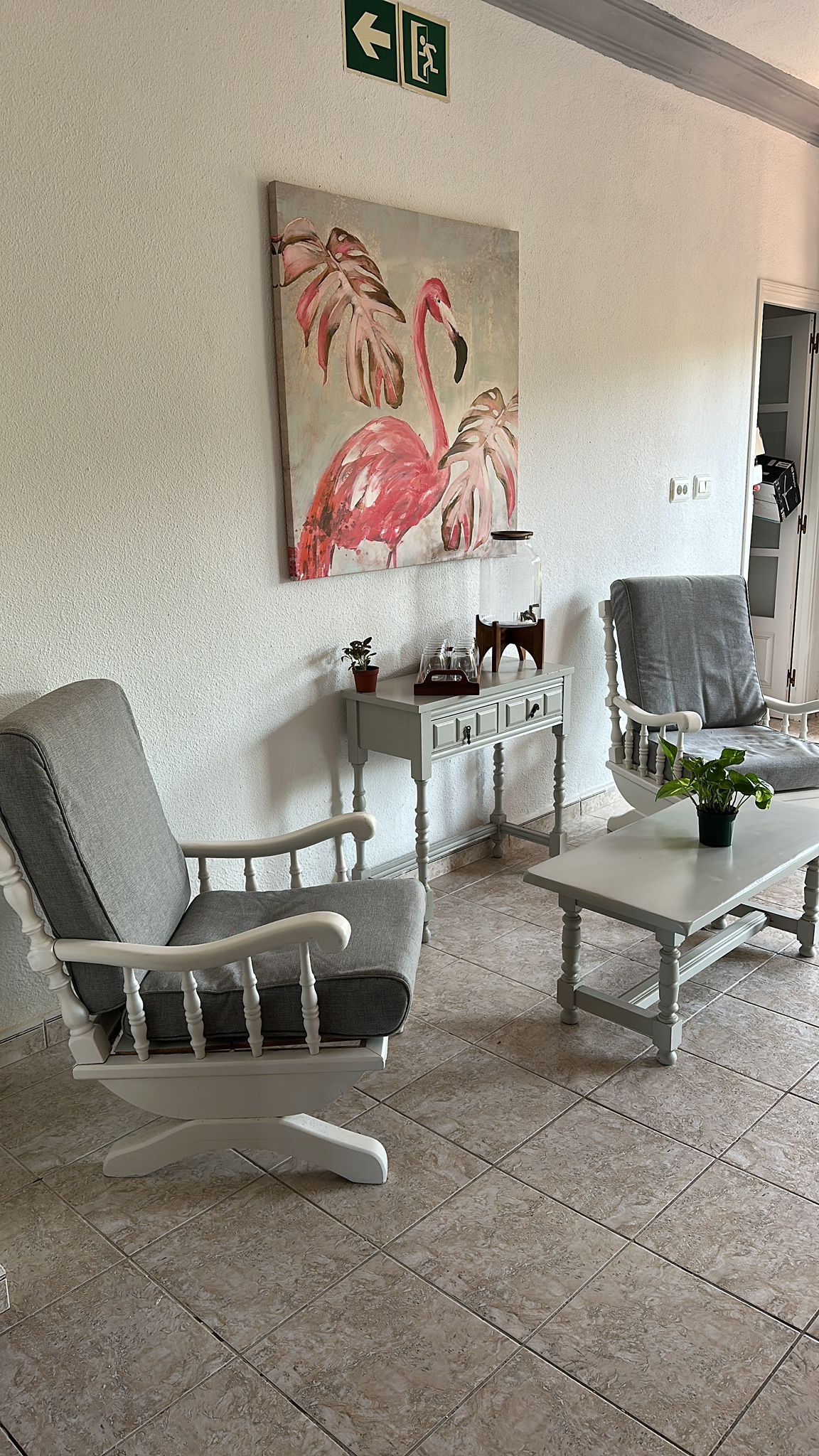 A room with a white chair, a table, a painting of flamingos, and a small plant.