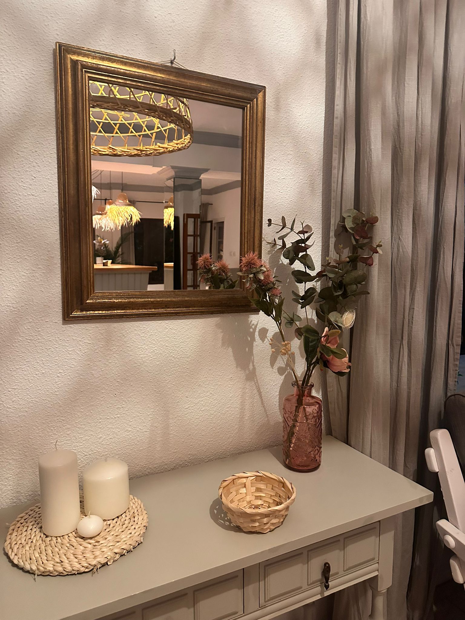Une pièce avec un miroir, un vase rempli de fleurs, des bougies et un panier en osier posé sur une commode.