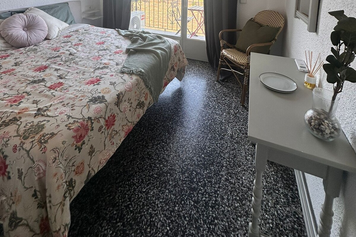 A hotel room with a bed, a chair, a table, and a view of the outside.