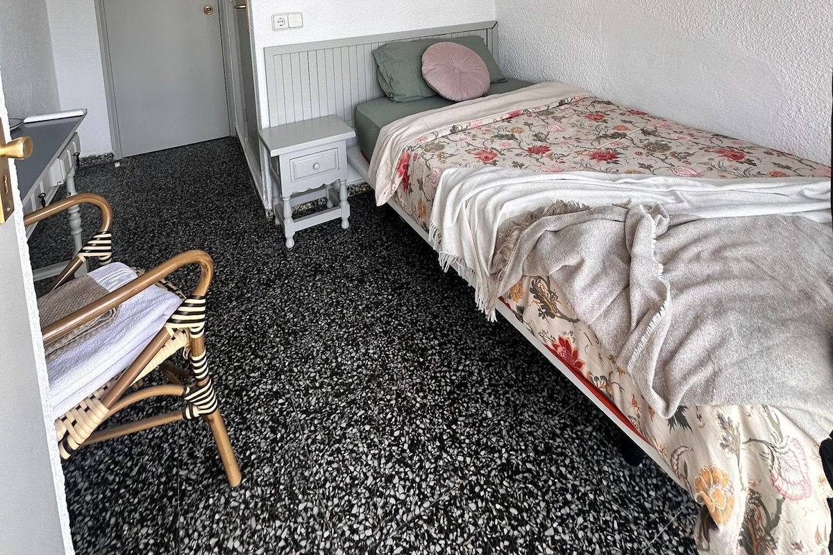 Habitación pequeña y funcional con una cama individual, una mesita de noche, una silla y una puerta que da a otra estancia.