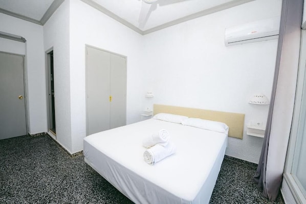 A hotel room with a bed, a door, an air conditioning unit, and a wall-mounted shelf.