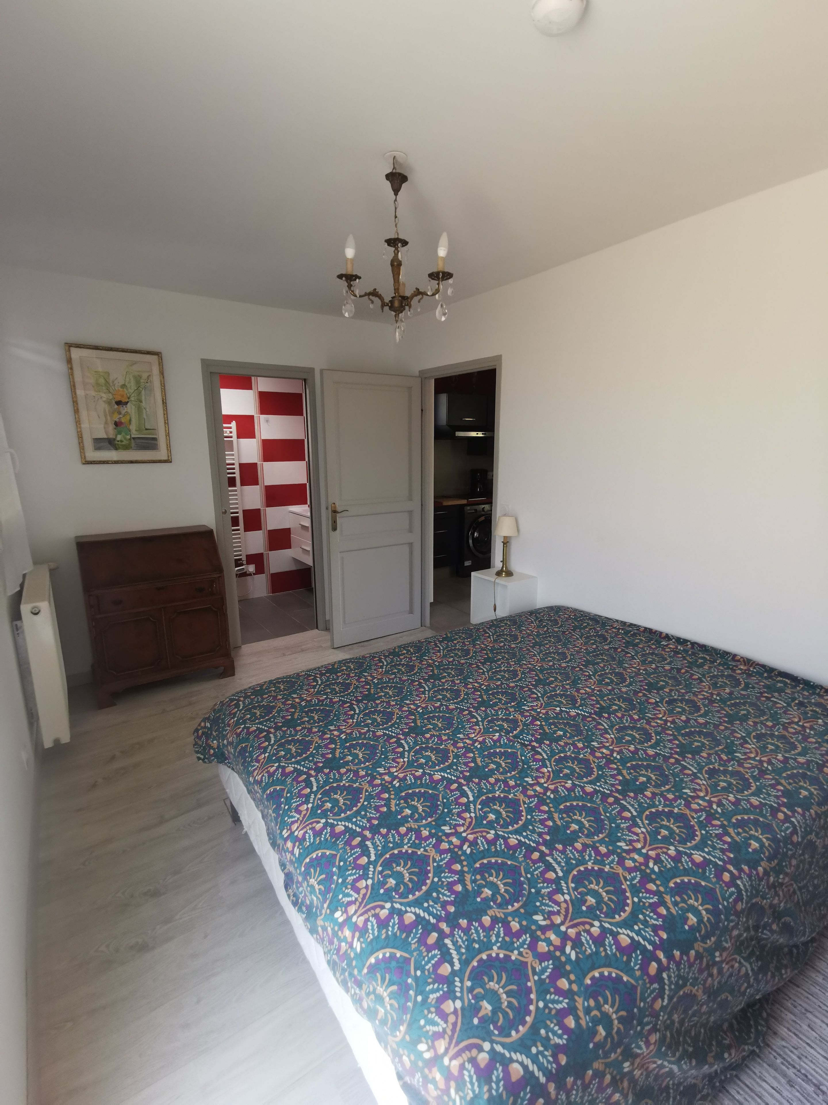 A bedroom with a patterned bedspread, a wooden dresser, a chandelier, and a door leading to a laundry area.