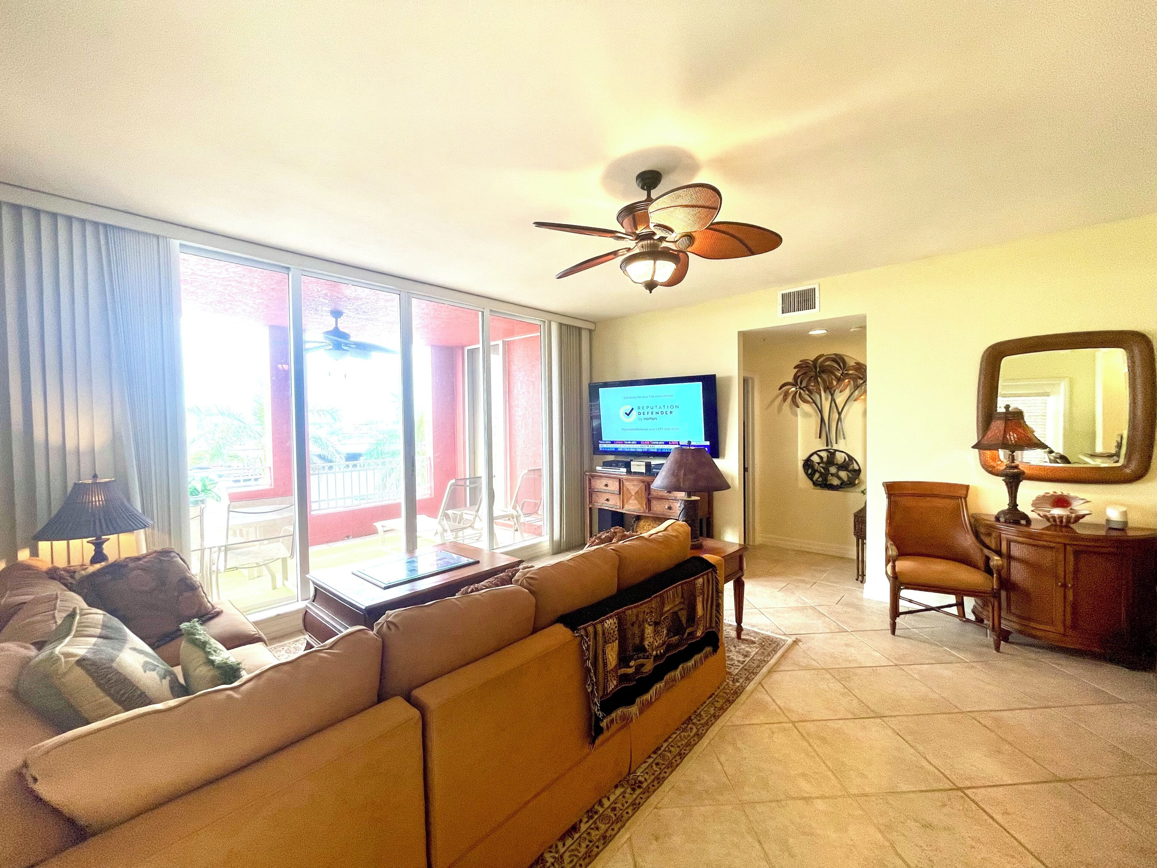 A living room with a sofa, a television, a ceiling fan, and a view through large windows.