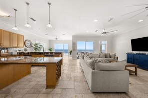 A spacious living area with a large window overlooking the ocean, a kitchen with wooden cabinets, and a ceiling fan. - Hammock Dunes East (Orange Beach)