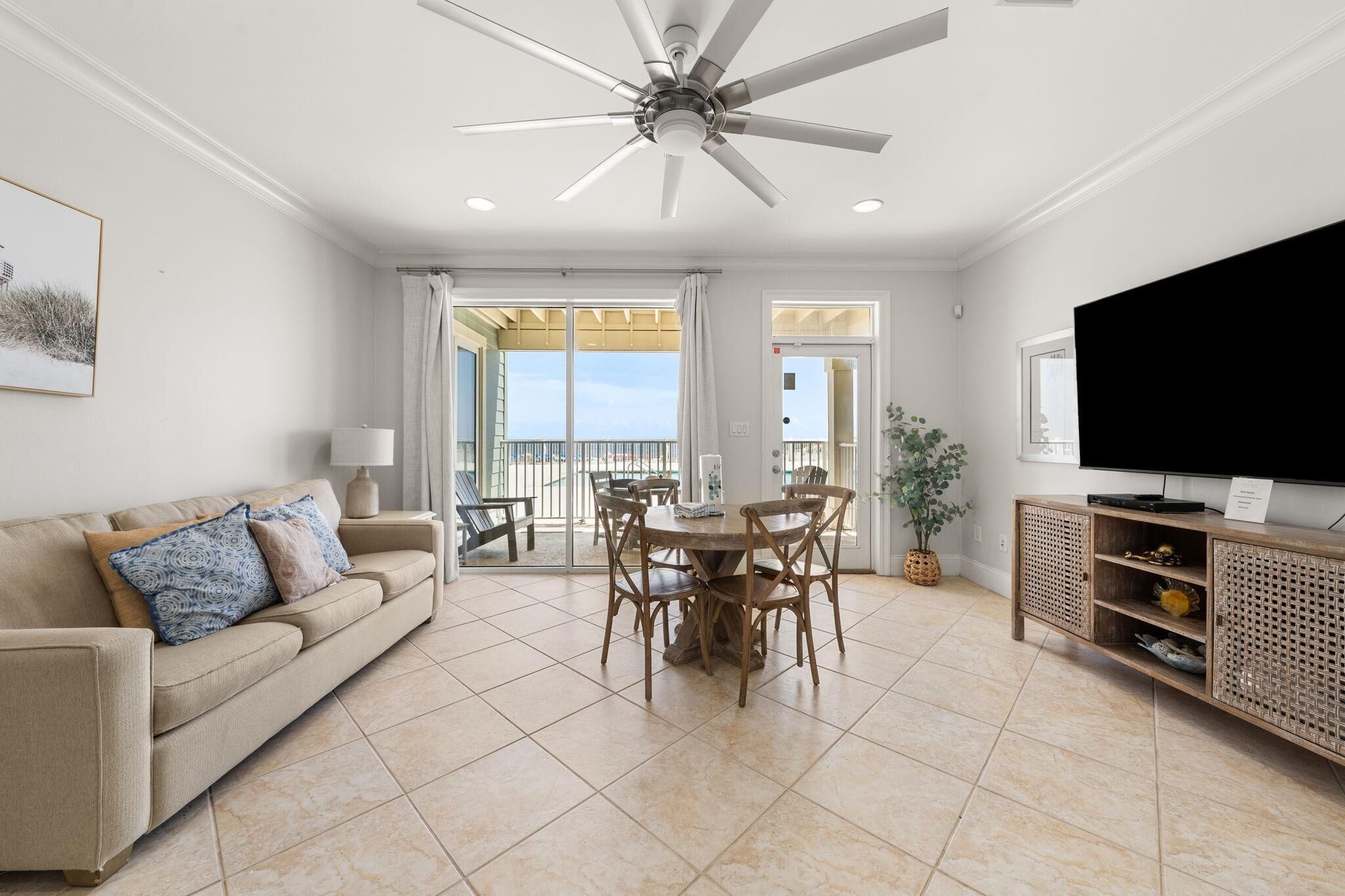 A spacious living room with a ceiling fan, a large flat-screen TV, a dining area with wooden chairs, and a balcony with a view.