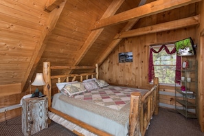 A wooden cabin bedroom with a bed, a nightstand, a lamp, a clock, and a window with curtains. - Critically Acclaimed Views Of The Smokies (PIGEON FORGE)