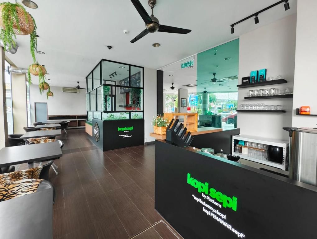 Un área de recepción moderna con un mostrador negro que muestra el nombre 'kopisepti' y una partición de vidrio. Hay zonas de estar con mesas y sillas, y se puede ver un ventilador de techo.