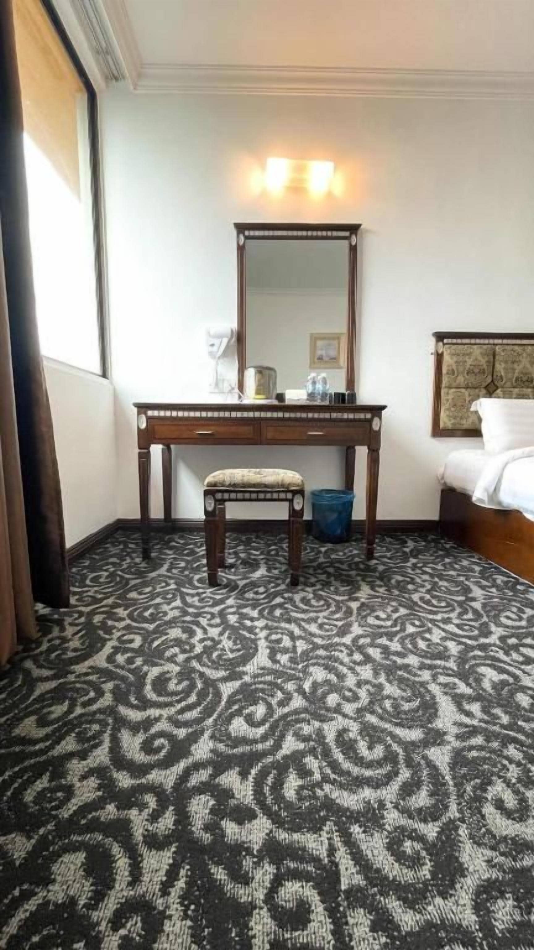 A hotel room with a bed, a desk with a chair, a large mirror, and a patterned carpet.
