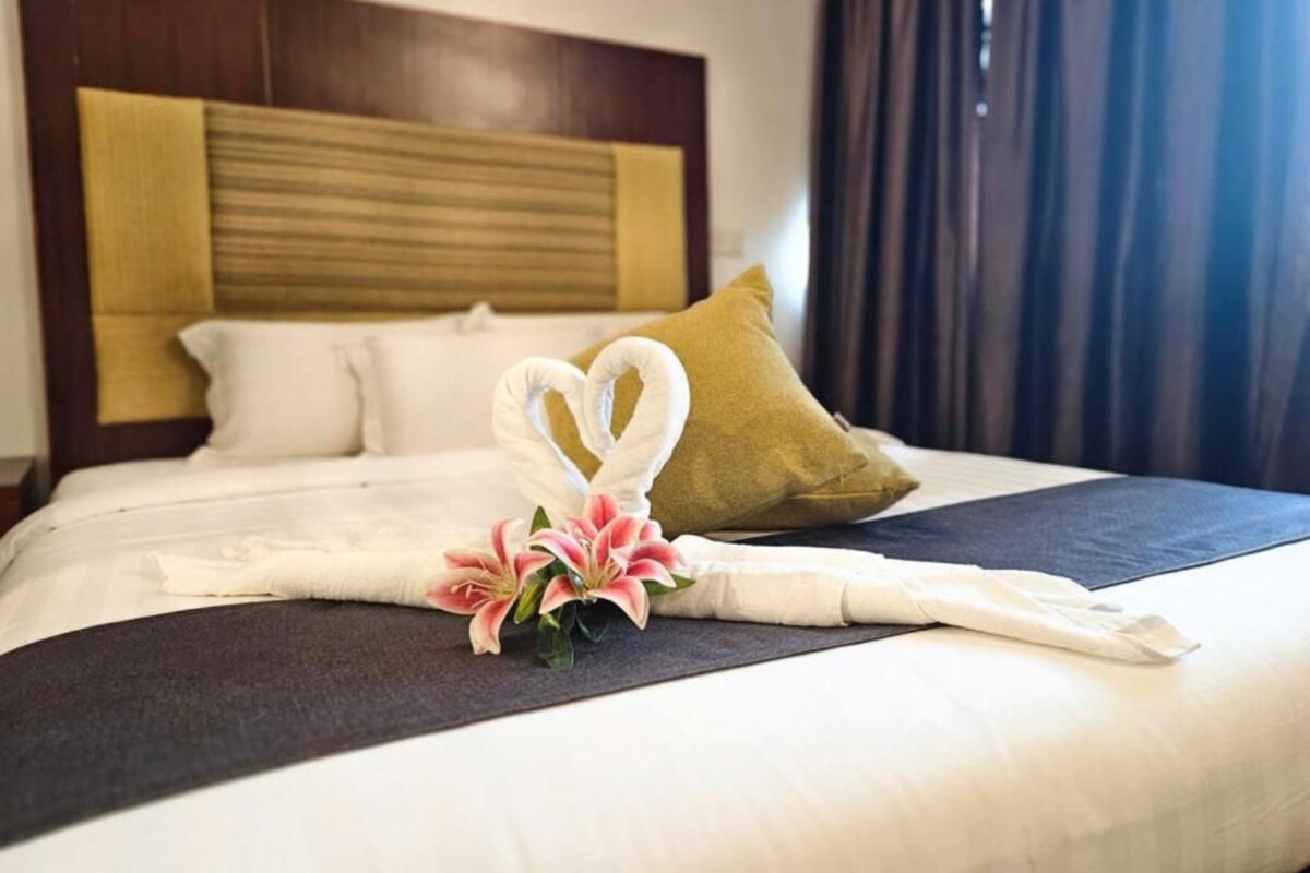A neatly made bed with a decorative towel and flowers, a wooden headboard, and a dark curtain.