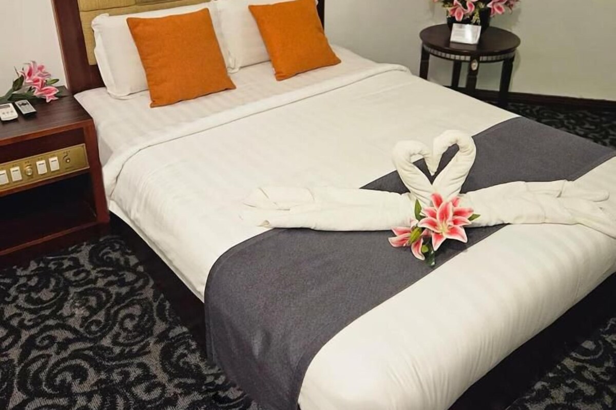 A hotel room with a bed, pillows, a bedside table, a nightstand, and a flower arrangement.