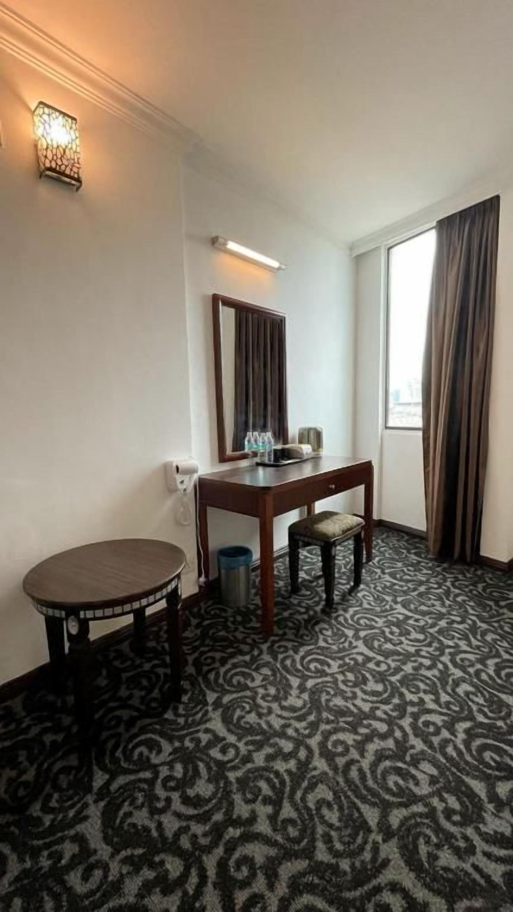 Een hotelkamer met een bed, een klein tafeltje, een stoel, een bureau, een spiegel, een lamp en een raam met gordijnen.