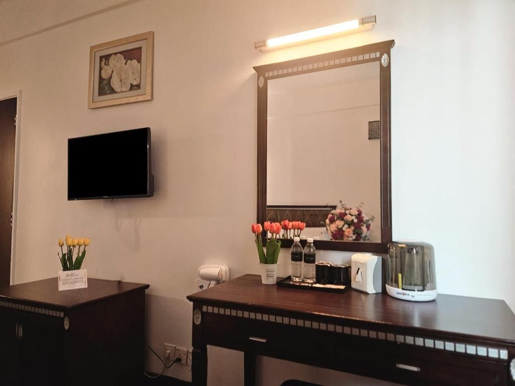 A hotel room with a flat-screen TV mounted on the wall, a wooden desk with a mirror, a toaster, and a vase of flowers.