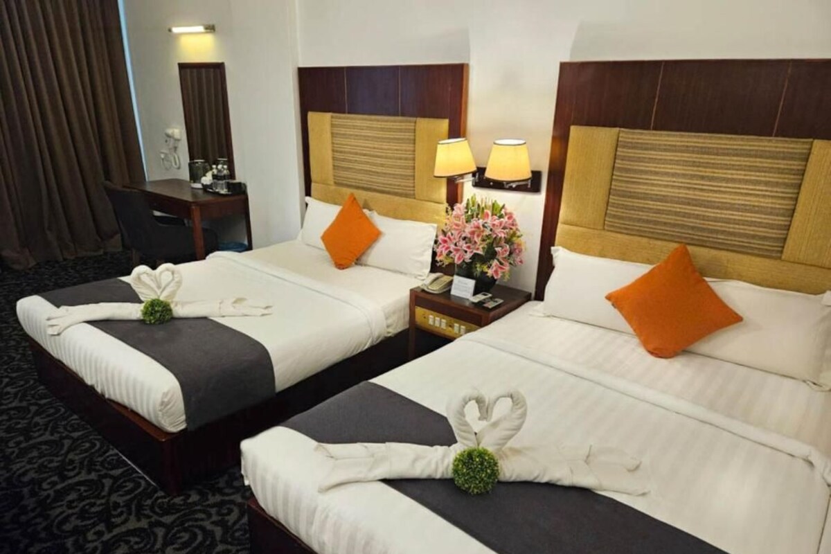 Two hotel beds with towels arranged in a heart shape, a small table with a lamp, and a desk with a chair.
