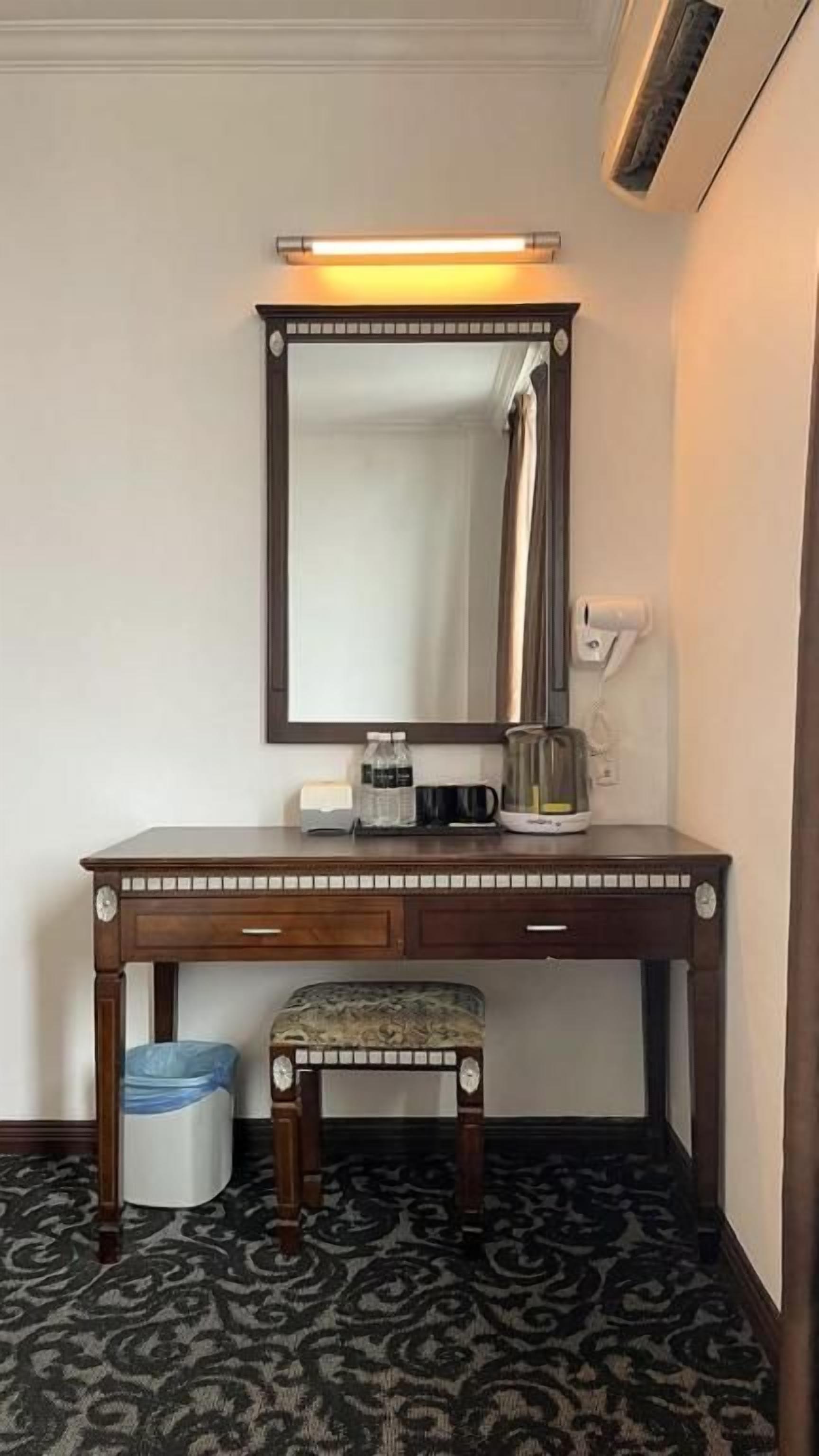 A hotel room with a wooden vanity, a mirror, a small bench, a trash bin, and a patterned carpet.