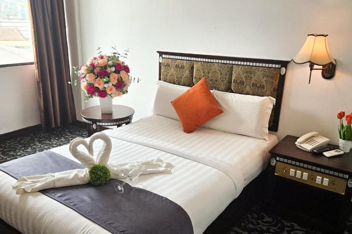 A hotel room with a neatly made bed, a bedside table with a lamp, and a floral arrangement on the nightstand.