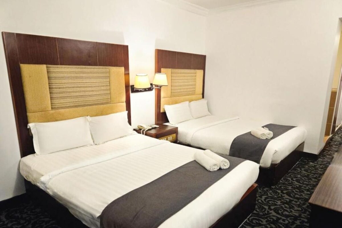 A hotel room with two beds, each with a wooden headboard, white bedding, and a small bedside table with a lamp.