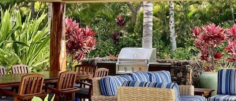 Un espace extérieur aménagé en terrasse, avec un barbecue, des meubles en osier et des plantes tropicales.