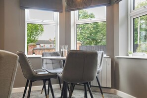 A corner room with two grey chairs, a small table, and a view of a brick building through large windows. - The Olive Tree Aparthotel | Deluxe Single Stay (Kingston upon Hull)