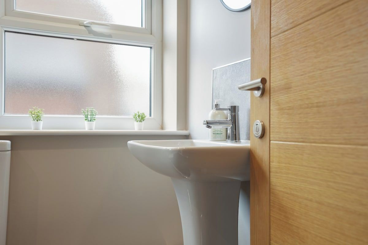 Ein Badezimmer mit einem weißen Waschbecken, einer Holztür und einem Fenster mit zwei kleinen Pflanzen auf dem Fensterbrett.