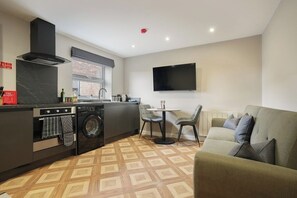 A compact living space with a kitchenette, a washing machine, a dining area with chairs, and a flat-screen TV mounted on the wall. - The Olive Tree | Deluxe 2-Bed Apartment (Kingston upon Hull)