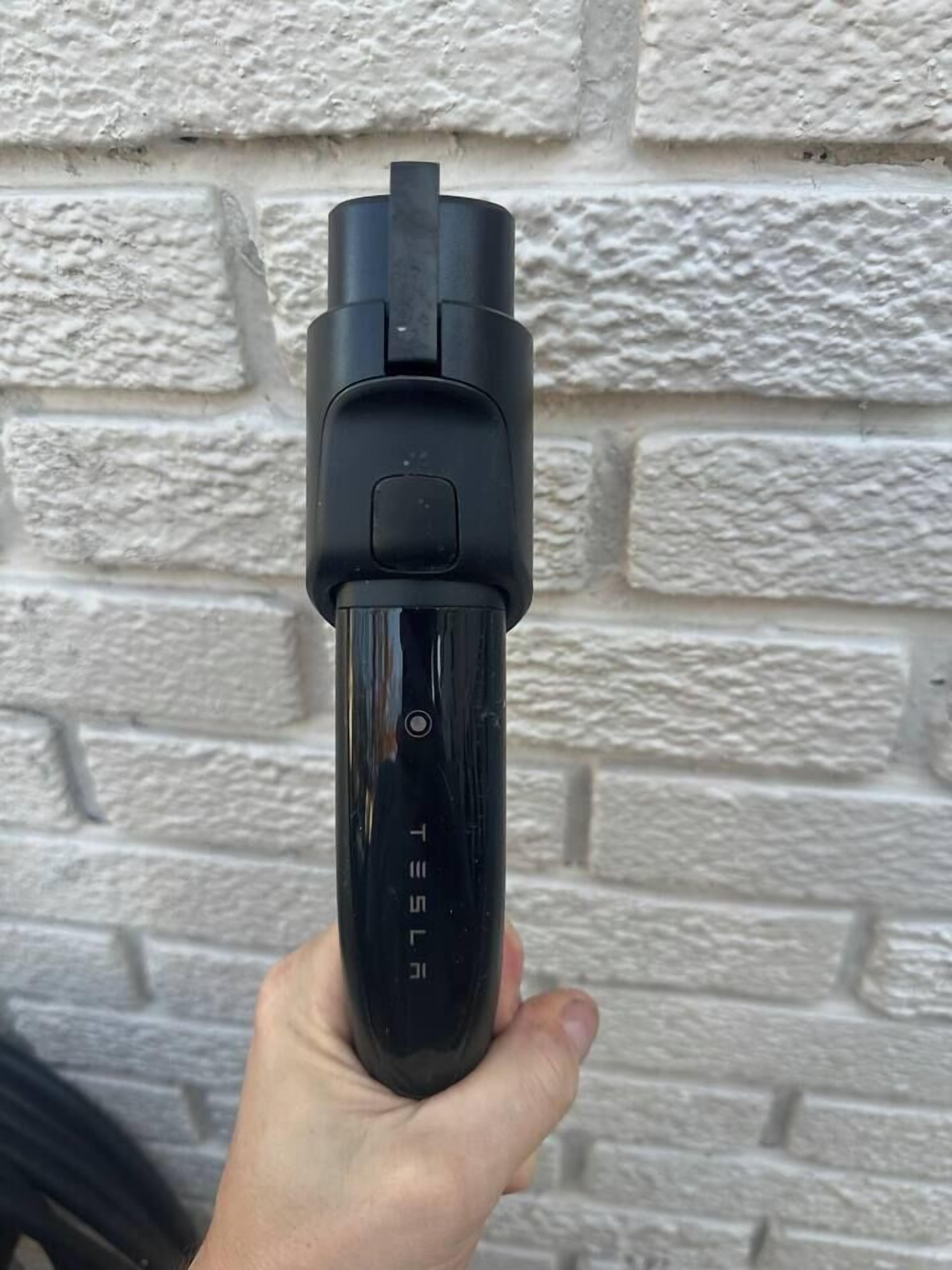 Una mano sosteniendo un objeto negro con un botón y una pantalla, sobre un muro de ladrillos blancos.