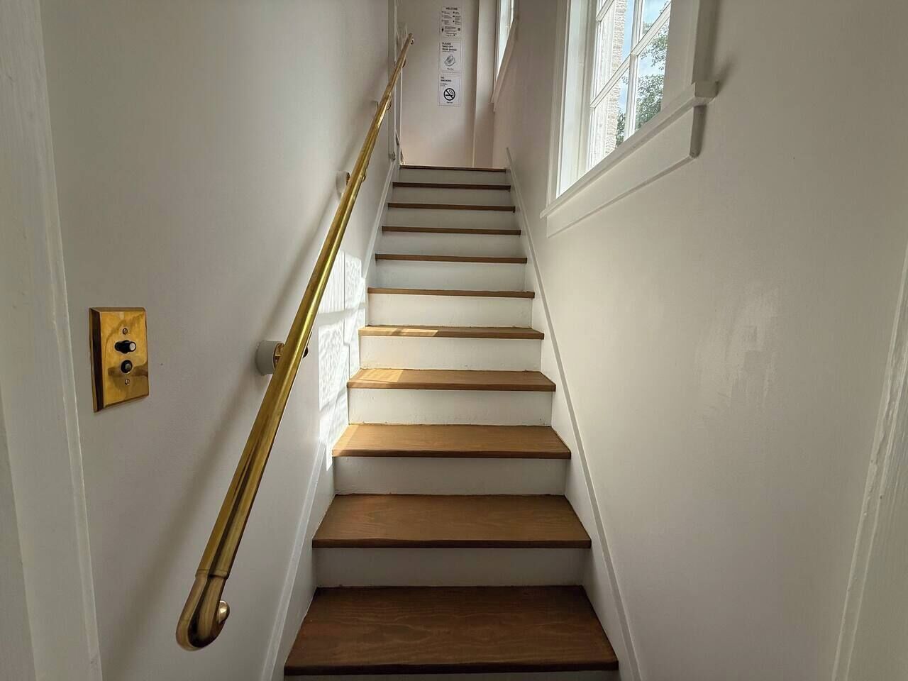 A staircase with wooden steps and a brass handrail, leading to a landing with a door and a light switch.