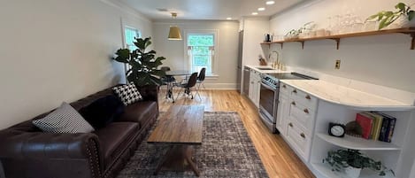 A modern kitchen with white cabinetry, a dining area with a brown leather couch, and a large window providing natural light.
