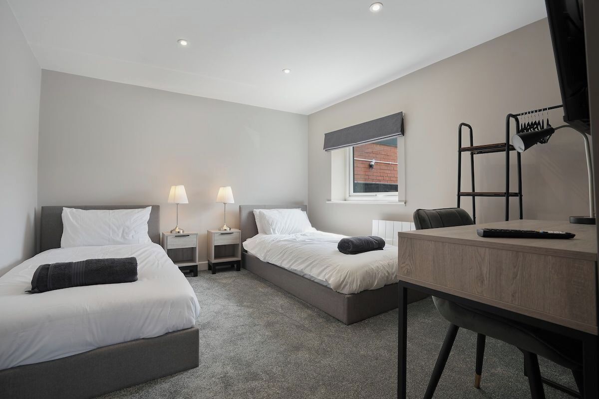 A hotel room with two beds, a desk, a chair, a window, and a lamp.