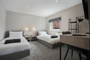 A hotel room with two beds, a desk, a chair, a window, and a lamp. - The Olive Tree | Cozy Deluxe 2-Bed (Kingston upon Hull)