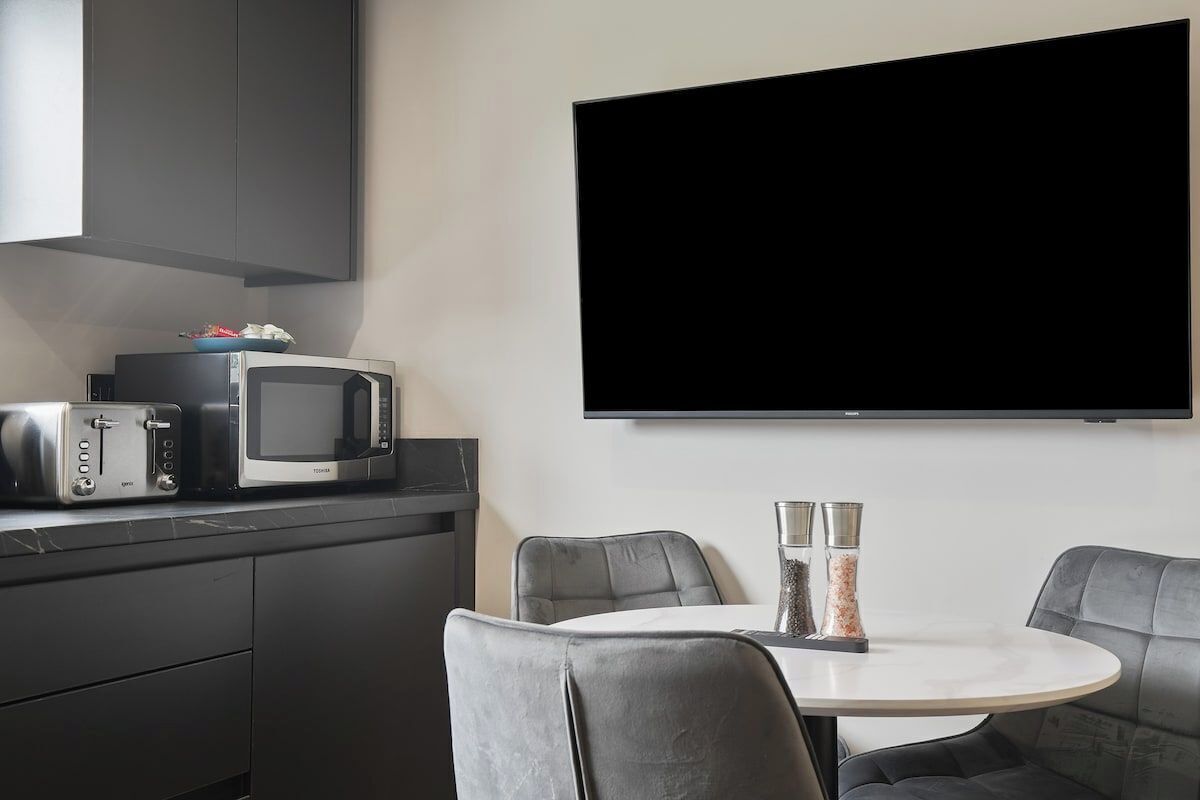 A modern kitchen with a flat-screen TV, a microwave, and a toaster. There is a round table with two chairs and two salt shakers on the table.